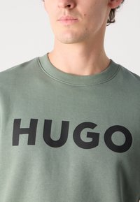 Olivengrønn sweatshirt laget av bomullsmiks. Har rund hals og stor svart tekst som sier "HUGO" over framsiden.