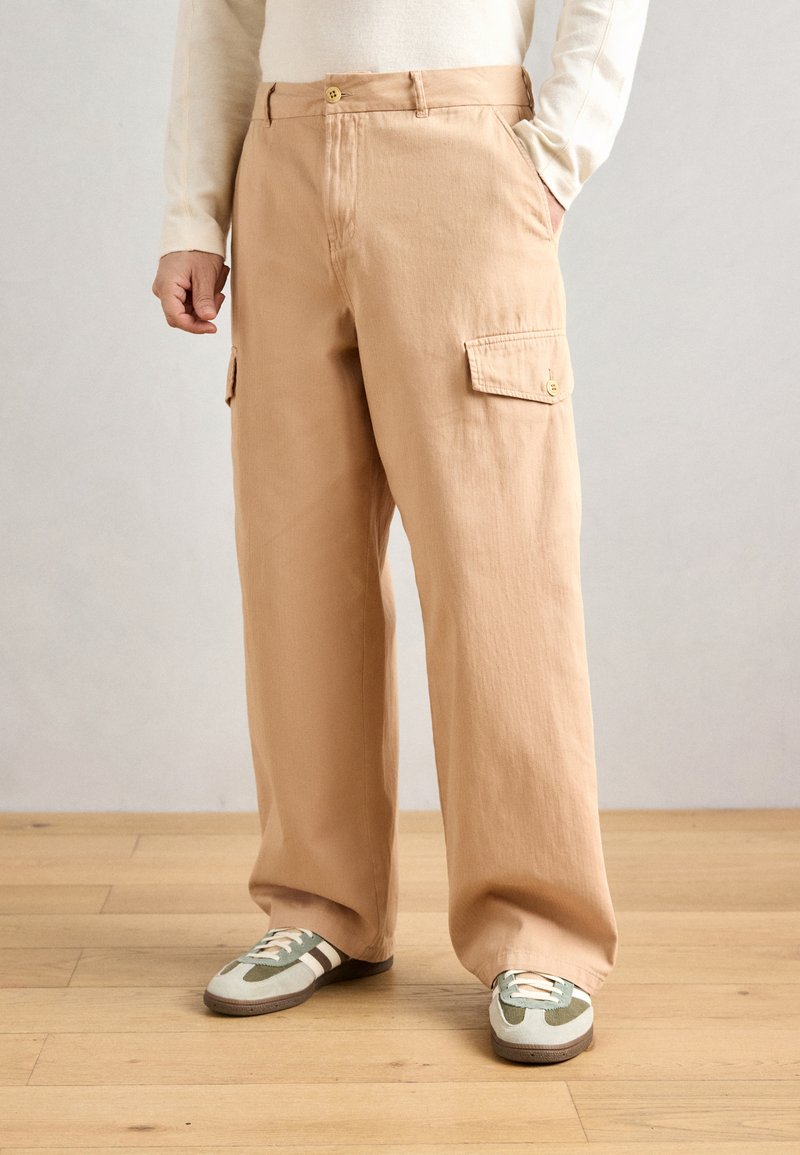 Beige cargo pants with a relaxed fit, featuring two side pockets and a single back pocket, made from soft fabric. Paired with green-accented sneakers.