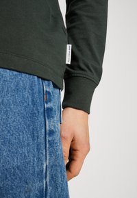 Camisa de manga larga de color verde oscuro con puños acanalados, combinada con unos vaqueros de mezclilla azul claro que presentan áreas desgastadas y un pequeño detalle de etiqueta naranja.