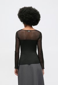 Moteris su garbanotais plaukais, vilkinti juodus permatomus ilgais rankovėmis marškinius ir pilką sijoną, pavaizduota iš nugaros prieš paprastą baltą foną.