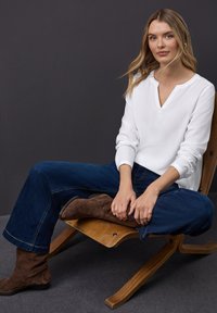 Blusa bianca a scollo a V con maniche lunghe, abbinata a jeans svasati blu scuro e stivali in pelle scamosciata marrone. Seduta su una sedia di legno contro uno sfondo scuro.