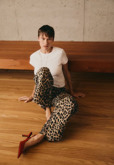 White fitted t-shirt, leopard print wide-leg pants, red stiletto heels. Wooden bench and floor; gray textured wall as backdrop.