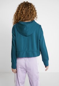 Haut à capuche teal, coupé, avec une capuche, présentant des manches longues et des poignets côtelés. Porté avec un pantalon violet clair avec des boutons-pression sur le côté.