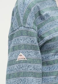 Φλις πουλόβερ σε μπλε χρώμα με πράσινη και μπλε ριγέ σχέδιο. Περιλαμβάνει μια εντυπωσιακή τριγωνική εφαρμογή λογότυπου που αναγράφει "Quiksilver" στο μανίκι.