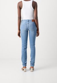 Mujer vestida con jeans ajustados de color azul claro, top sin mangas blanco y sandalias de tacón beige, de pie frente a un fondo blanco liso.