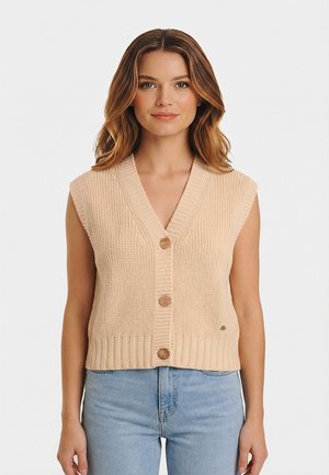 Femme portant un gilet beige en tricot sans manches avec boutons et un jean taille haute bleu clair, debout devant un fond uni.