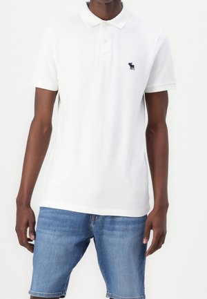 Biała koszulka polo z krótkim rękawem wykonana z bawełny, z trzema guzikami i małym granatowym logo na piersi, zestawiona z dżinsowymi szortami.