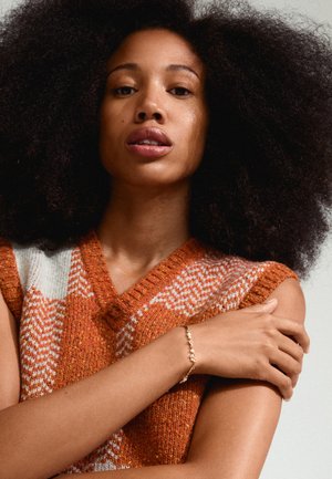 Maglione lavorato a maglia in arancione e crema con scollo a V; modello che indossa un braccialetto con piccole perle. I capelli sono voluminosi e ricci.