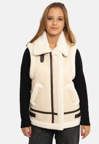 Jeune femme portant un gilet sans manches en peau retournée crème avec un grand col, par-dessus un haut noir à manches longues, debout devant un fond uni.