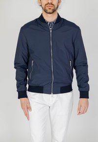 Giacca bomber navy in tessuto leggero con zip frontale, due tasche laterali, polsini e cintura a coste. Indossata sopra una camicia a fantasia.