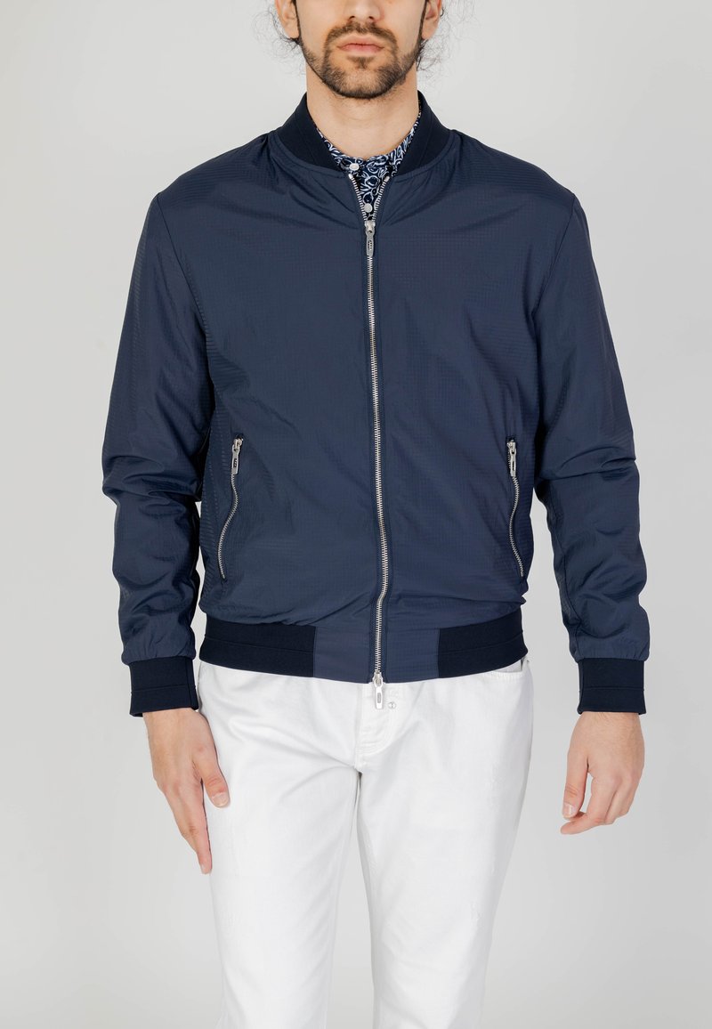 Giacca bomber navy in tessuto leggero con zip frontale, due tasche laterali, polsini e cintura a coste. Indossata sopra una camicia a fantasia.