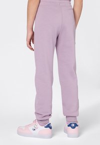 Ein Kind in lila Jogginghosen und weiß-rosa-blauen Turnschuhen steht vor einem weißen Hintergrund, von hinten betrachtet.
