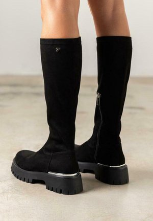 Botas de ante negras hasta la rodilla con una suela negra gruesa y un acento plateado. Cuentan con una cremallera lateral y una textura suave.