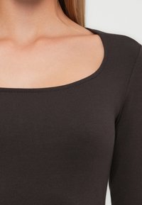 Nærbilde av en person som har på seg en mørkebrun genser med rund hals og lange ermer, som viser utsnittet ved halsen, skulderen og øvre del av brystet.