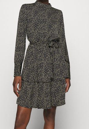 Czarna sukienka na długi rękaw w białe kropki, z wysokim dekoltem, wiązaniem w talii i wielowarstwową spódnicą, noszona przez osobę stojącą na jednolitym tle.