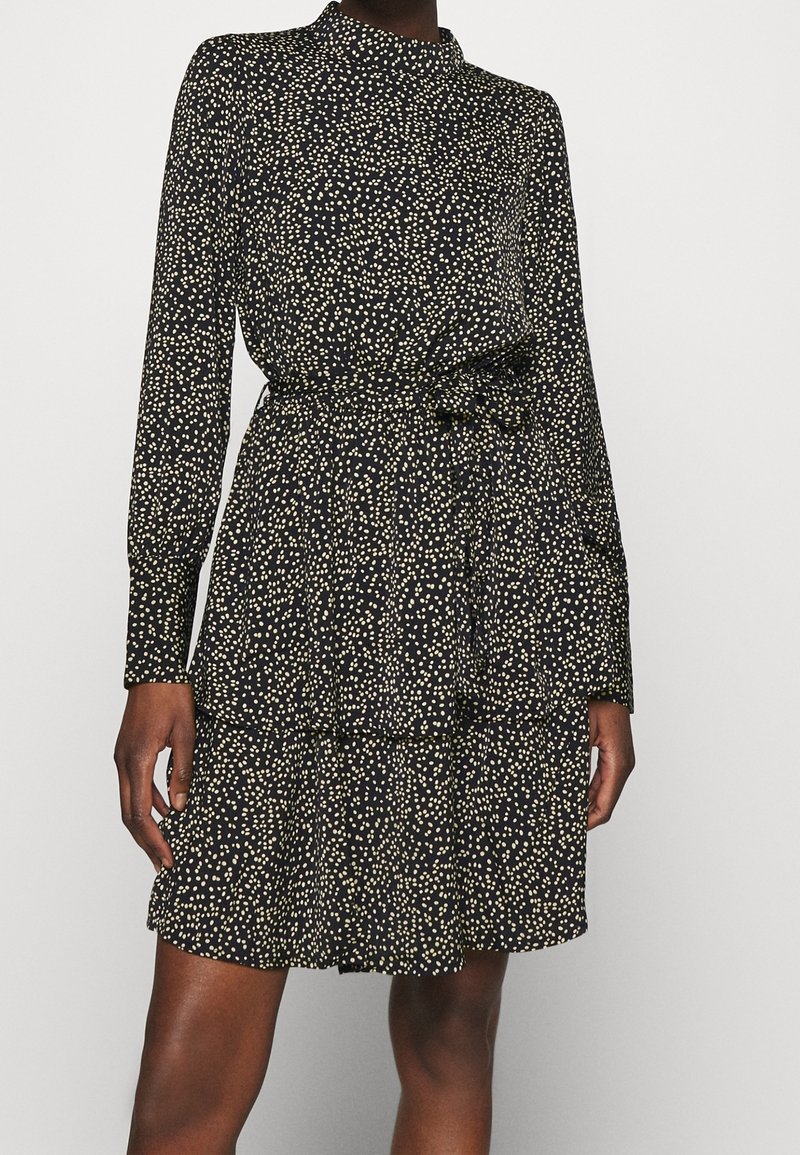 Robe noire à manches longues avec pois blancs, col haut, ceinture à la taille et jupe à volants portée par une personne debout devant un fond uni.