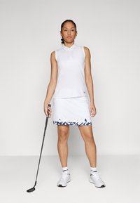 White sleeveless polo shirt paired with a white golf skirt featuring a navy abstract pattern at the hem. The model holds a golf club.