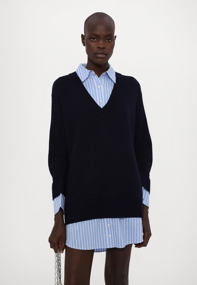 Marineblauer Strickpullover mit tiefem V-Ausschnitt, über ein hellblaues, gestreiftes Hemd mit Knöpfen gelegt. Verfügt über überschnittene Schultern und gerippte Bündchen.