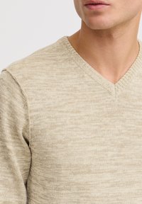 Beige neulepusero V-pääntiellä, joka esittelee teksturoitua kangasta vaaleassa, vaihtelevassa kuosissa. Kappale on lähellä kehoa istuva.