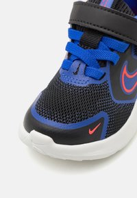 Chaussure de sport noire et bleue avec tige en mesh, accents réfléchissants et semelle blanche. Équipée d'une sangle noire et de lacets bleus pour un maintien sécurisé.