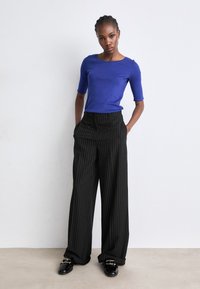 Blue fitted shirt with short sleeves paired with wide black pinstriped trousers. Model stands with hands in pockets, wearing shiny black shoes.