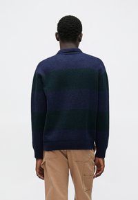 Tengerészkék és zöld csíkos pulóver bordázott gallérral és mandzsettával. Puha kötött anyagú, kényelmes fazonú, a hátsó részen látható varrási részletekkel.