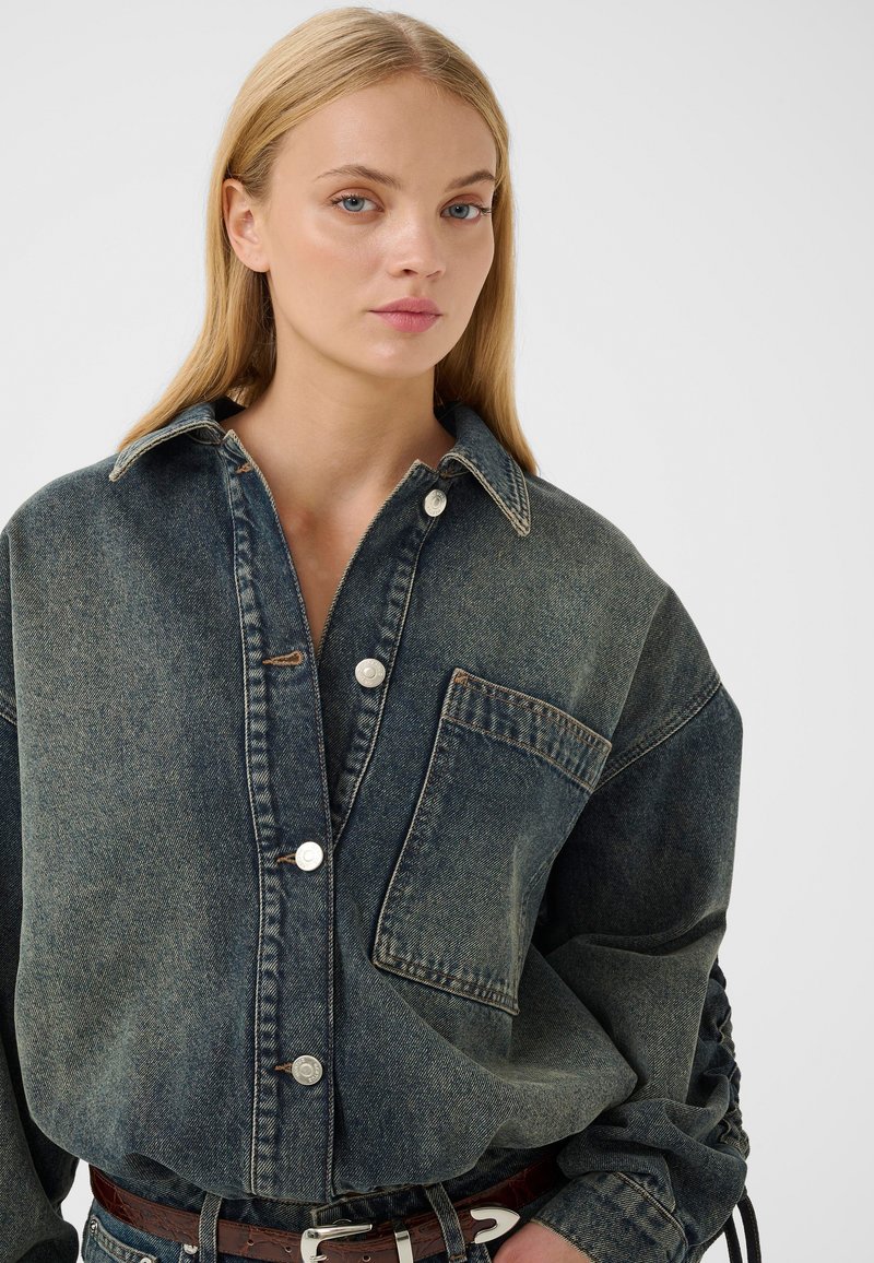 Jeune femme portant une chemise oversize en denim foncé avec une grande poche avant et des boutons argentés, debout devant un fond clair uni.
