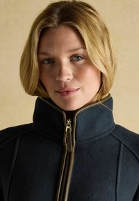 Tmavomodrá fleecová bunda s vysokým golierom, spevnená zlatým zipsom a hnedými detailmi pozdĺž okrajov. Hladká textúra a priliehavý strih.