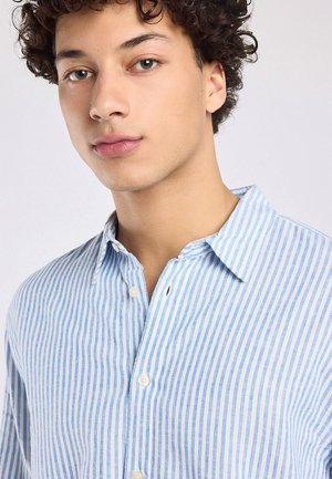 Jonge man met krullend haar die een lichtblauw-wit verticaal gestreept overhemd draagt, iets naar links gedraaid.