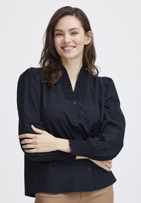 Čierna blúzka s gombíkmi a textúrovaným výstrihom, s nafúknutými rukávmi, vyrobená z hladkej tkaniny s priliehavým pásom.