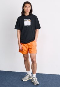 Svart T-skjorte med grafisk trykk og tekst, sammen med oransje shorts. Utseendet fullføres med grå og hvite sneakers.
