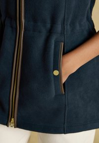 Tengerészeti fleece dzseki arany cipzárral és barna díszítéssel. Elöl zsebe van, amely patentos részlettel rendelkezik. Puha tapintás, testreszabott szabás.