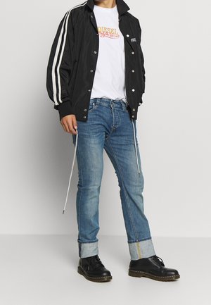 Veste en nylon noire avec des rayures blanches, t-shirt blanc avec un graphique coloré, jeans bleu clair et bottes noires à lacets avec une semelle robuste.