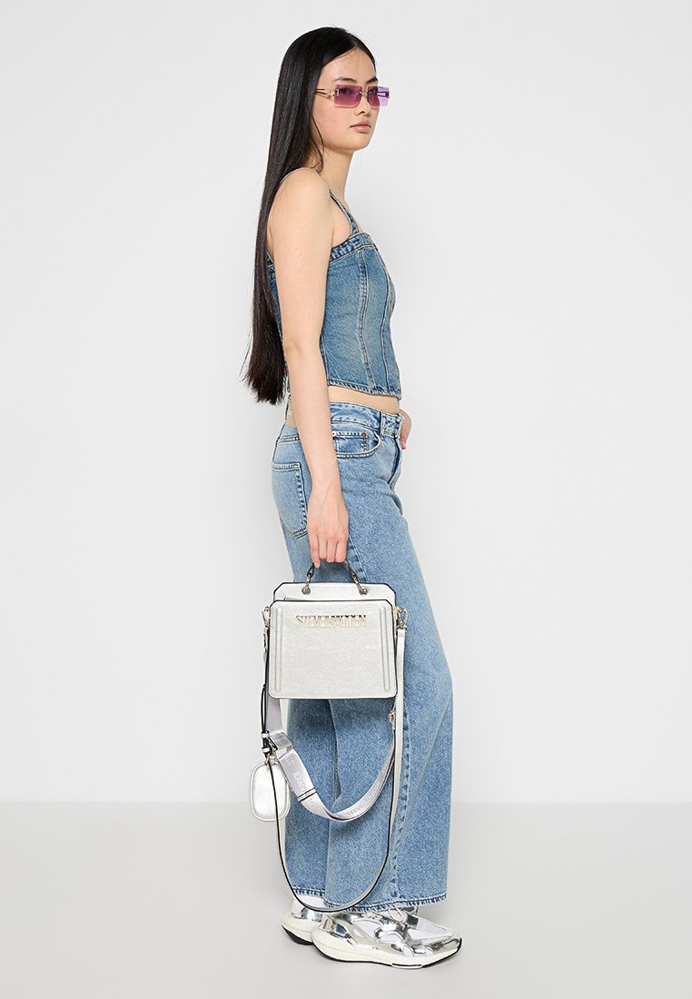 Sac à main argenté avec poignées supérieures et bandoulière. Présente un logo embossé, une forme structurée et une finition métallique, associé à une tenue en denim.