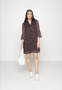 Vestido preto floral com mangas transparentes em forma de sino, apresentando um decote em V e um corte solto. O modelo segura uma bolsa branca texturizada e usa sapatos brancos.