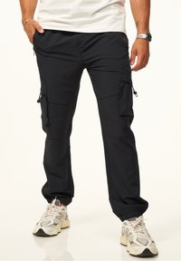 Schwarze Cargo-Hosen mit elastischen Bündchen, mehreren mit Reißverschluss und Knopf verschlossenen Taschen sowie einer eleganten, glatten Stoffoberfläche. Kombiniert mit hellen Sneakers.