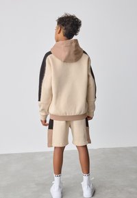 Beige hættetrøje med brune og sorte detaljer, kombineret med matchende beige shorts. Fremstillet af blødt stof, med rummelig hætte og ribstrikkede manchetter.