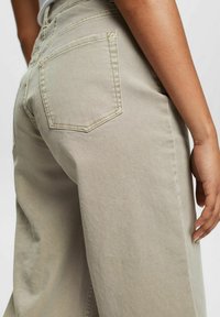 Jean beige clair à jambes larges avec une finition texturée, comportant une poche arrière et un détail de couture visible. Main reposant sur la hanche.
