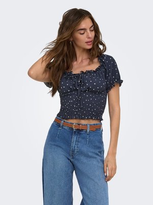 Kvinde med langt brunt hår iført en marineblå stump top med polkaprikker og højtaljede blå jeans med et brunt bælte, poserer med den ene hånd i håret.