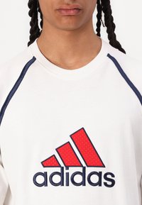 Hvid sweatshirt med et rødt adidas-logo og diagonale detaljer. Blå piping langs skuldrene giver kontrast til det enkle design.