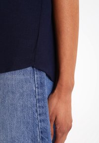 Top a tinta blu navy, con texture e orlo curvo, abbinato a jeans in denim chiaro. Primo piano di una mano appoggiata sul tessuto dei jeans.