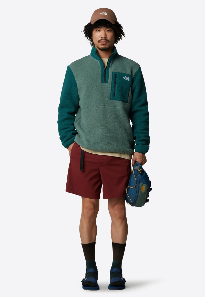 Polarowy półgolf w kolorze teal i ciemnej zieleni, z zippowanym kieszenią na piersi. Połączony z bordowymi szortami i niebieską torbą na ramię. Brązowa czapka na głowie.