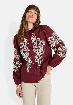 Chemise boutonnée bordeaux avec broderie blanche détaillée sur les manches, col volanté et manches bouffantes ; dotée de boutons en bois.