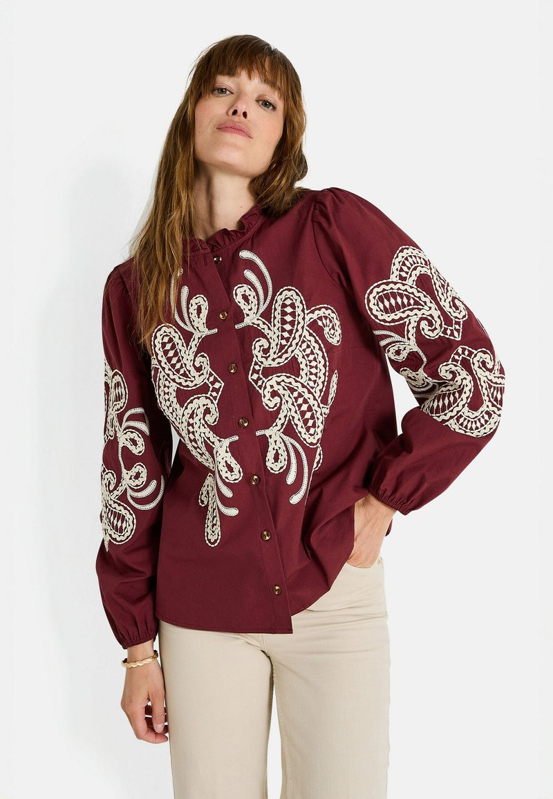 Chemise boutonnée bordeaux avec broderie blanche détaillée sur les manches, col volanté et manches bouffantes ; dotée de boutons en bois.