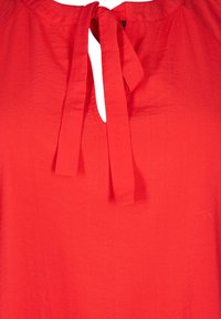 Blusa rossa con scollatura a goccia e dettaglio legato, realizzata in tessuto leggero con una consistenza morbida e un leggero luccichio.