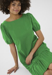 Zelena kratkoročnu obleko s plisiranim robom, izdelano iz gladkega materiala. Ima okroglo ovratnik in naberene rokave za dodaten detajl.