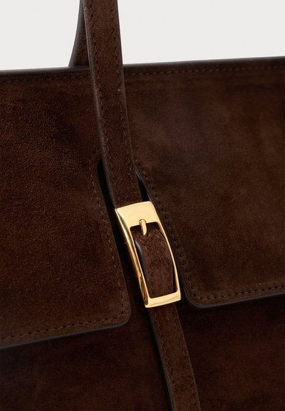Κοντινή λήψη σε τσάντα χειρός από σκουρόχρωμο καφέ σουέντ με ραμμένο καπάκι και λεπτό λουράκι που ασφαλίζει μια γυαλιστερή χρυσή ορθογώνια αγκράφα.