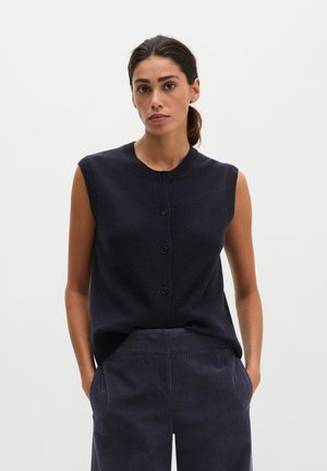 Woman with dark hair in a low ponytail wears a sleeveless dark buttoned vest and dark pinstriped pants, standing with hands in pockets.