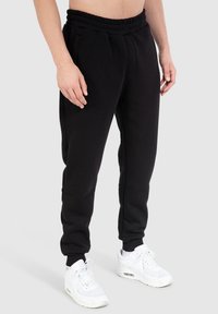 Schwarze Sweatpants mit elastischem Bund, seitlichen Taschen, eng zulaufendem Bein-Design und gerippten Bündchen, kombiniert mit weißen Sportschuhen.