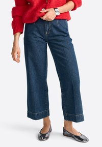 Mujer vestida con pantalones de mezclilla recortados de pierna ancha, suéter de punto rojo, bailarinas metálicas y una llamativa pulsera plateada en la muñeca.